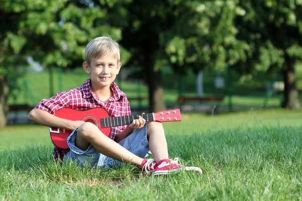 Niño feliz sentado en la hierba y tocar la guitarra Imagen de archivo