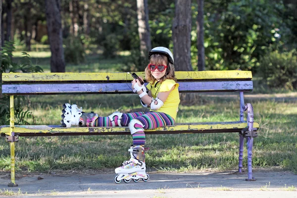 Holčička s kolečkové brusle sedí na lavičce a naslouchat mu — Stock fotografie