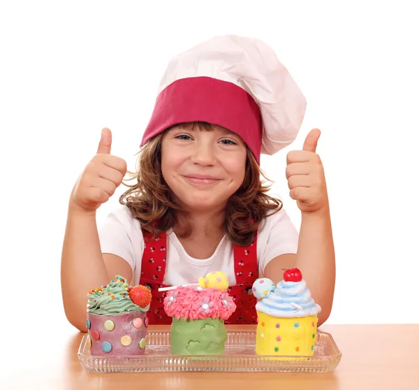 Happy little girl cook with cupcakes and thumbs up — Stock Photo, Image