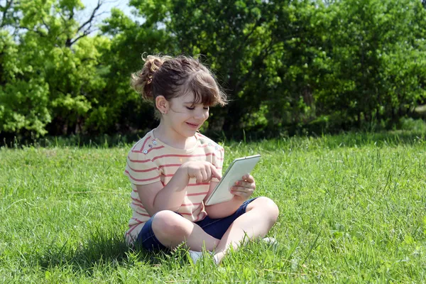 Happy little girl sitting on grass and play with tablet — Stock Photo, Image