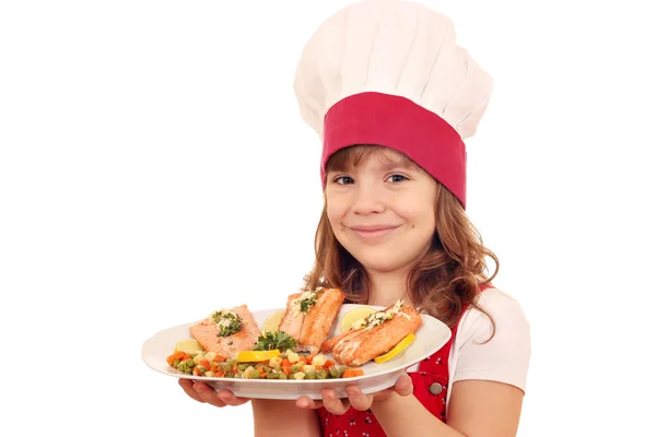 Heureuse petite fille cuisinier avec du saumon et des légumes sur le plat — Photo