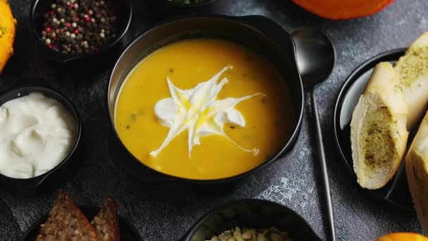 Deliciosa sopa de calabaza con crema, semillas, pan y hierbas frescas en un elegante tazón negro de cerámica — Vídeos de Stock