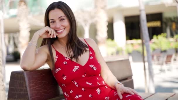 Mujer morena hispana sonriente con un vestido rojo se sienta en un banco en el parque — Vídeo de stock