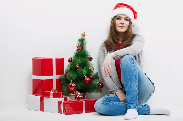 Vrouw in Kerstman hoed zitten in de buurt van de kerstboom en geschenken. — Stockfoto