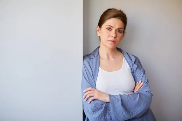 Woman with serious face wearing blue pajamas standing near gray wall.