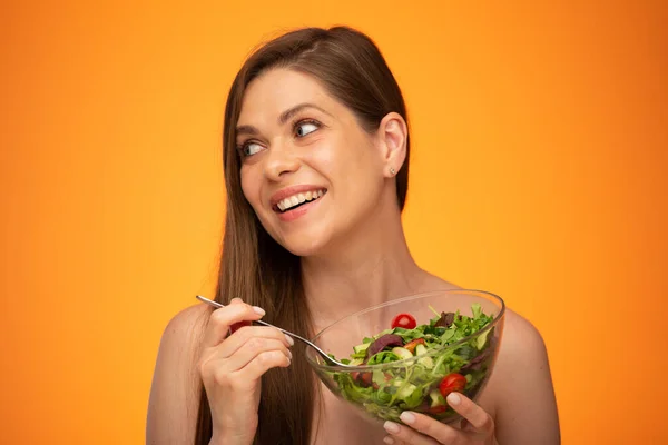 Laughing woman with bared shoulders holding green salad in glass bowl and looking away isolated portrait over orange yellow background.