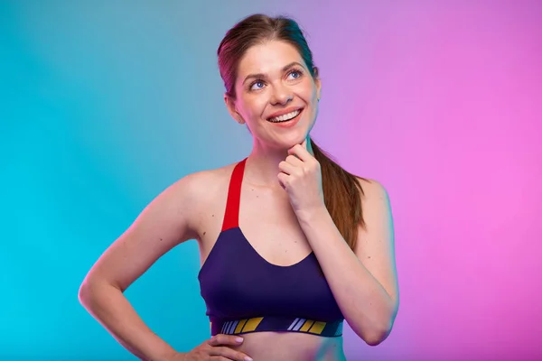 Thinking smiling sporty woman in fitness bra sportswear looking up. Female fitness portrait isolated on neon multicolor background. Girl face with hand touching chin.