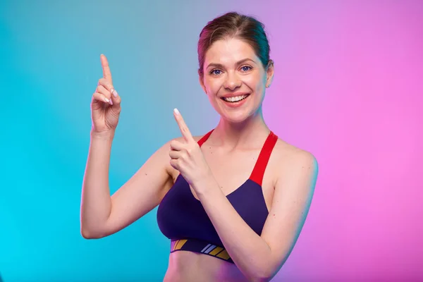 Smiling sporty woman in fitness sportswear pointing finger to empty space. Female fitness portrait isolated on neon multicolor background.