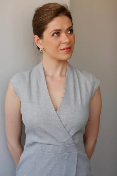 Smiling woman in gray dress holding hands behind back and look away.