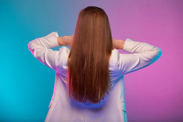 Woman with long hair standing back portrait with neon lights colors effect. Female model isolated on neon colored background wearing white suit.