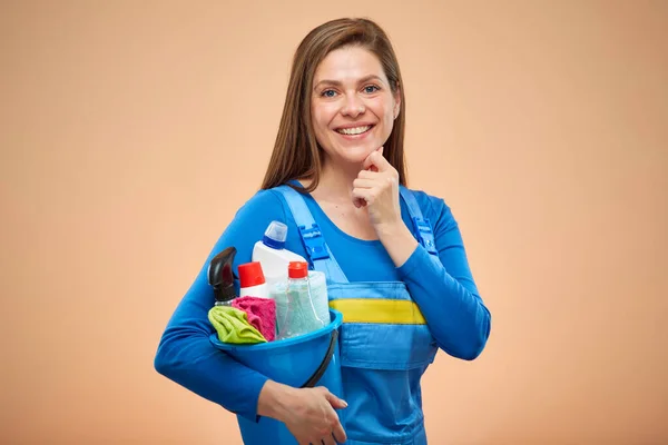 Thinking happy woman in overalls holding cleaning products in blue bucket. isolated on beige background.