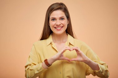 Gülümseyen kadın parmaklarıyla kalp hareketi yapıyor, sarı arka planda izole edilmiş bir portre.. 