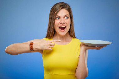 Gülümseyen ve boş tabağı parmağıyla gösteren bir kadın. Mavi arka planda izole edilmiş kadın portresi.