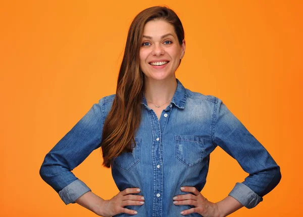 Smiling woman in blue denim shirt standing with hands on hips over orange background.