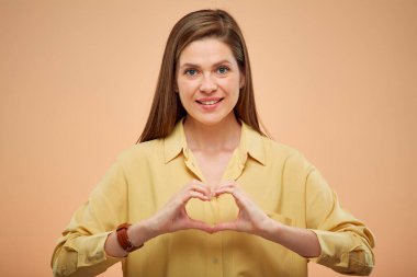 Gülümseyen genç bayan parmaklarıyla kalbi, stüdyo sarı arka planında izole edilmiş portresi.