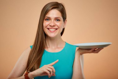 Elinde boş tabakla gülümseyen ve işaret eden kadın, sarı kahverengi arka planda izole edilmiş bir portre..