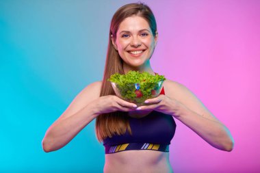 Spor kıyafetli, gülümseyen ve önünde cam kasede yeşil salata tutan bir kadın. Neon çok renkli arkaplanda izole edilmiş kadın fitness portresi.