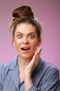 Şaşırtıcı bir şekilde pijamalı bir kadın elleriyle yüzüne dokunuyor, pembe renkte izole edilmiş, göz altı yamalı bir kadın yüzü..