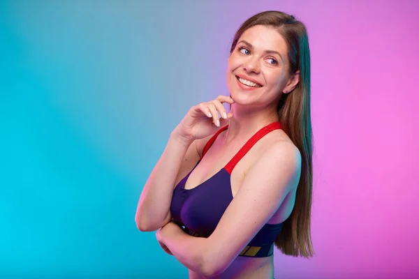 Thinking smiling sporty woman in fitness bra sportswear profile view, looking back over shoulder. Female fitness portrait isolated on neon multicolor background. Girl face with hand touching chin.