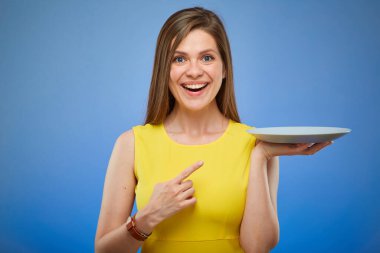 Gülümseyen ve boş tabağı parmağıyla gösteren bir kadın. Mavi arka planda izole edilmiş kadın portresi.