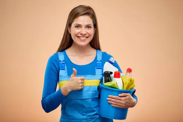 Cleaner lady in overalls holding bucket with cleaning products showing thumb up. isolated on beige background.