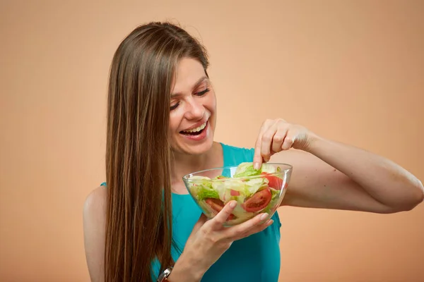 Young lady in azure dress eating green salad from glass bowl, isolated portrait on peach color background