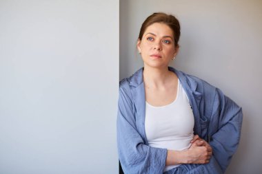 Ciddi yüzlü, mavi pijamalı bir kadın gri duvarın yanında duruyor..