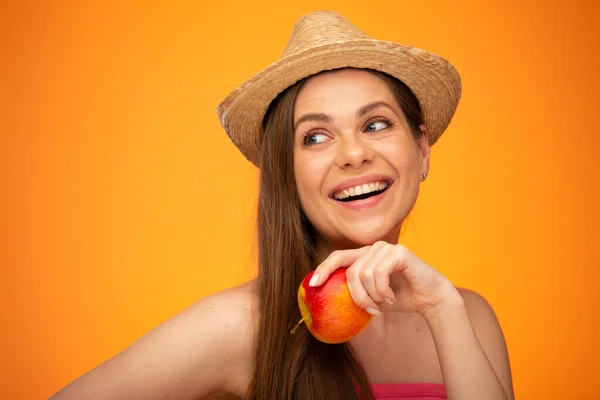 Happy woman with bared shoulders holding red apple and looking away over shoulder, face isolated portrait on orange background. Girl wearinh summer hat.