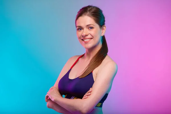 Smiling sporty woman in fitness sportswear with arms crossed. Confident female fitness coach portrait isolated on neon multicolor background.