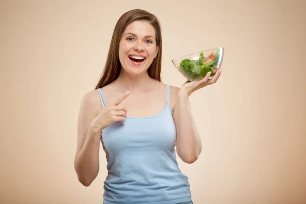 微笑的女人把绿色沙拉放在玻璃碗里 指指点点 在米黄色的背上挂着孤立的肖像 — 图库照片