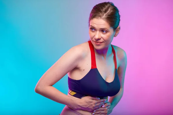 Woman looking back over shoulder, hand on stomach, abdominal pain and digestive disorder. sporty woman in fitness sportswear . Female fitness portrait isolated on neon multicolor background.