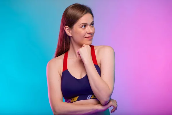 Thinking smiling sporty woman in fitness bra sportswear looking away. Female fitness portrait isolated on neon multicolor background. Girl face with hand touching chin.