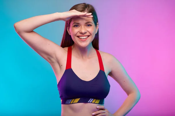 Sporty coach trainer woman in fitness sportswear looking for, with hand on forehead. Female fitness portrait isolated on neon multicolor background.