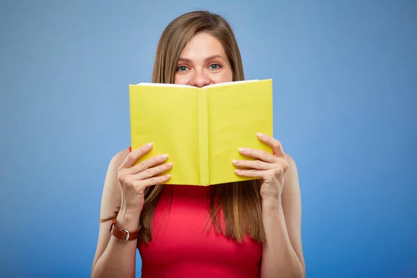 Student lady in red dress holding yellow book in front of face, isolated portrait on blue background.