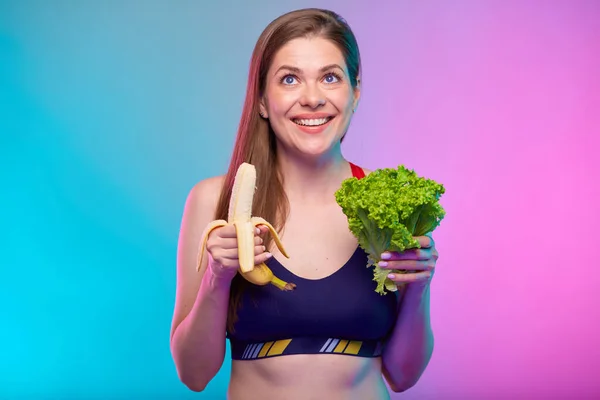 緑色のサラダ束とバナナを見上げて若い笑顔のスポーティな女性 健康食品とネオンカラーの背景に隔離された女性のフィットネスの肖像画 — ストック写真