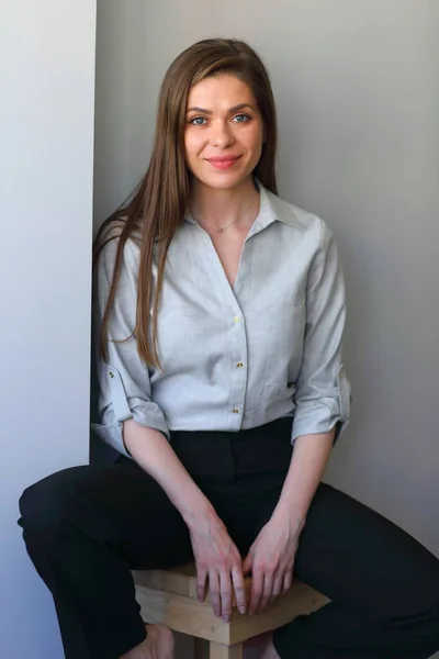 Smiling woman in blue shirt and black pants sitting on stool. Isolated portrait.