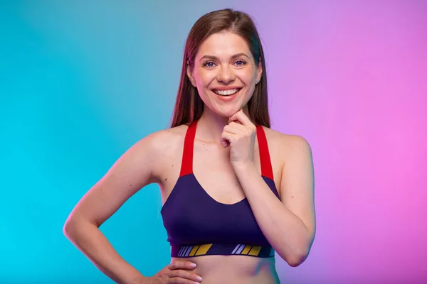 Thinking smiling sporty woman in fitness bra sportswear looking at camera. Female fitness portrait isolated on neon multicolor background. Girl face with hand touching chin.