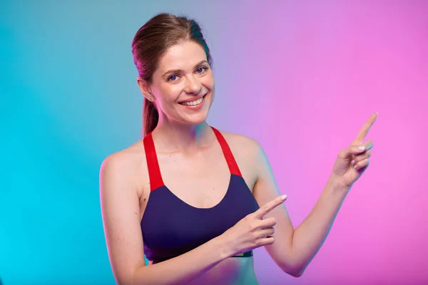 Sporty woman in fitness sportswear pointing finger up. Female fitness portrait isolated on neon color background.