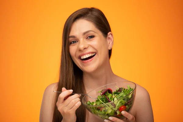 Smiling woman with naked shoulders holding bowl with green salad isolated on orange yellow background.