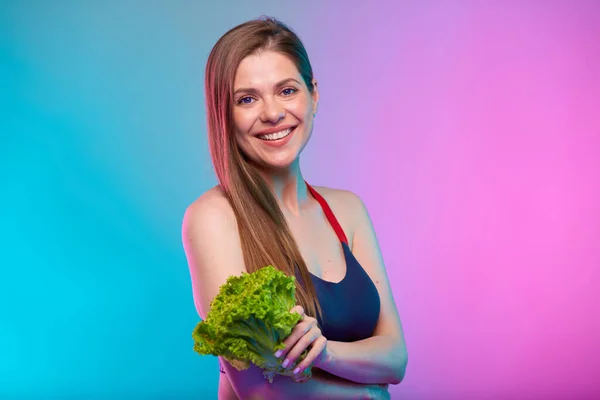 Smiling sporty woman in fitness bra sportswear holding green salad bunch. Female fitness portrait isolated on neon multicolor background.