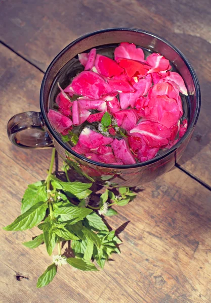 Het kopje thee met rozenblaadjes en mint op oude houten pagina — Stockfoto