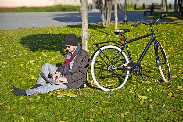 Un giovanotto che lavora su un portatile in un parco autunnale. La sua bicicletta st — Foto Stock