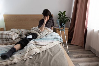 ACL ameliyatından sonra koltuk değnekleriyle yatakta oturan üzgün genç bir kadın..