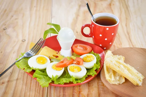Breakfast with hard boiled eggs, sliced in halves, cup of tea, salad, tomatoes, cheese and bread on the red plate and wooden background — Stock Photo, Image