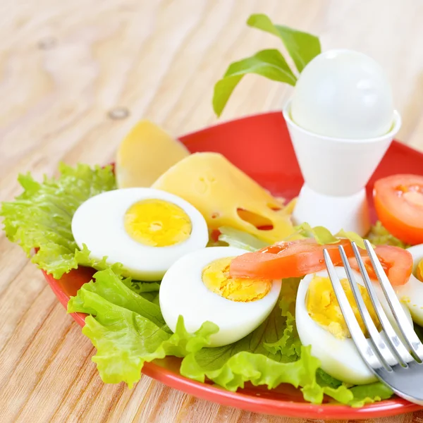 Ontbijt met hard gekookte eieren, gesneden in helften, salade, tomaten en kaas op de rode plaat en de houten achtergrond Stockfoto