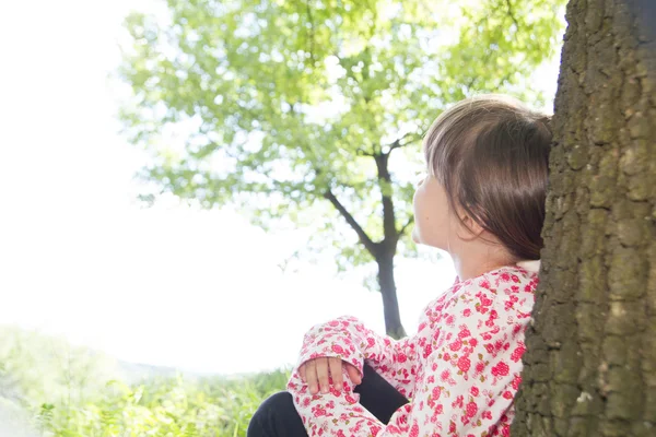 Маленькая милая девочка, прислонившаяся к дереву в лесу — стоковое фото