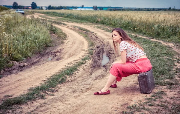 Krásná dívka čeká na venkovské silnici s starý kufr — Stock fotografie