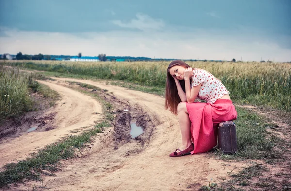 Hermosa chica esperando en un camino de campo con una vieja maleta — Foto de Stock