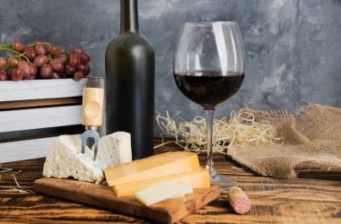 Kırmızı şarap tozlu bir şişe ve bardakta. Tahta bir masada üzüm ve peynir. Mesaj için yer var. Gri arkaplan.