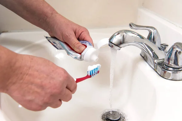 高齢女性の手は歯ブラシに歯磨き粉を適用します 水は開いているタップから注いでいます — ストック写真